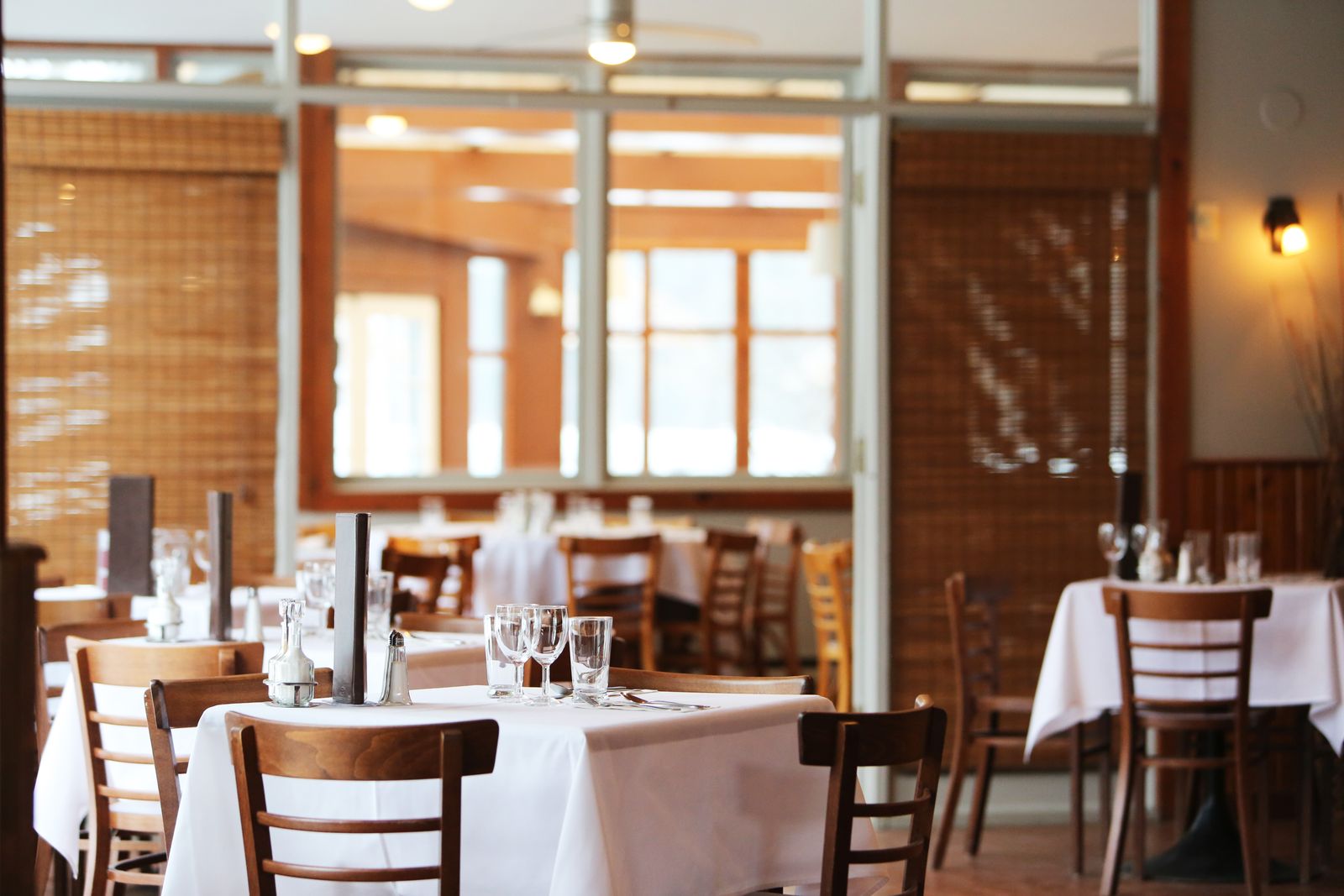 An empty restaurant dining room in daylight — white-linen tables set with glassware, wooden chairs, and bamboo blinds at the windows.