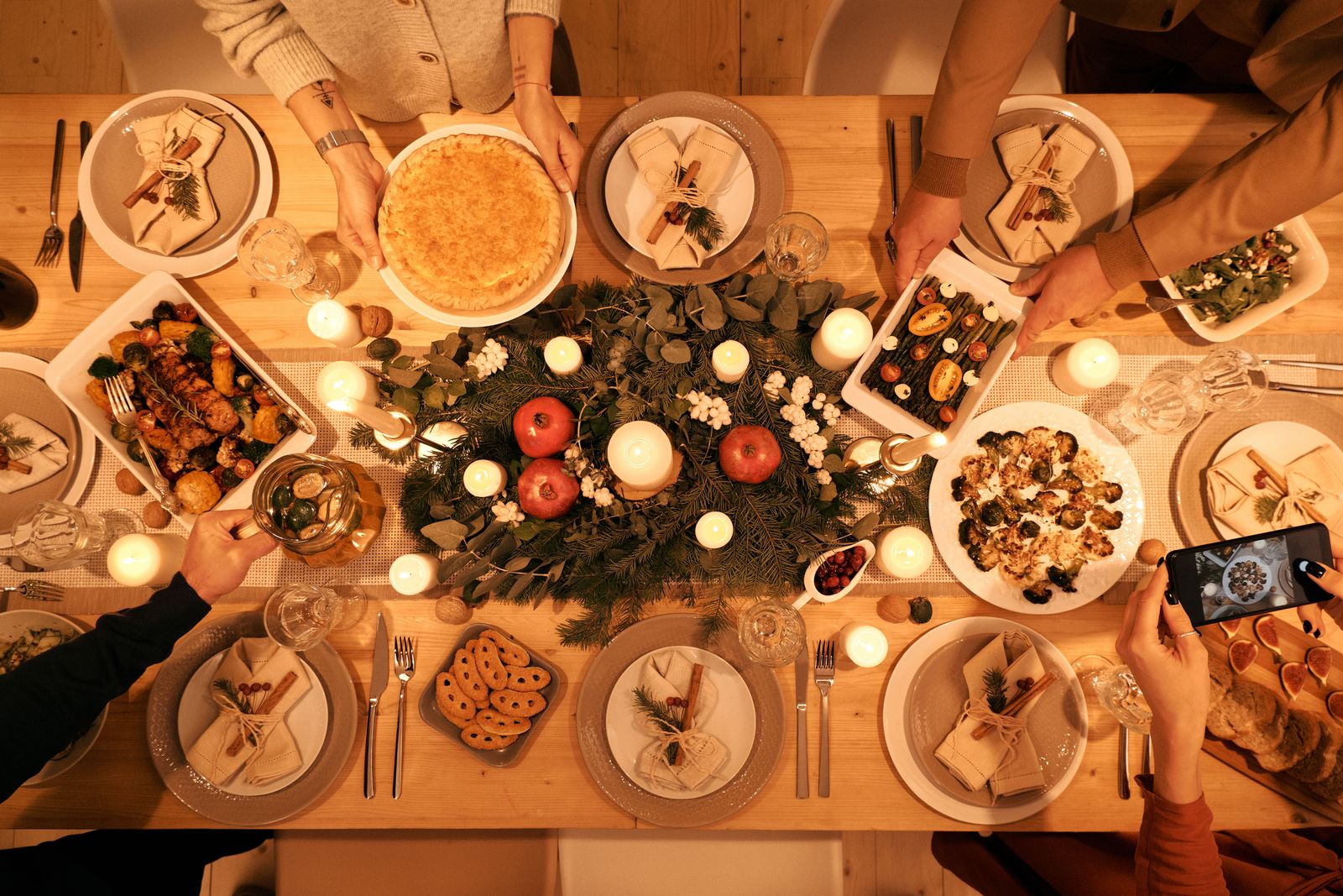 Overhead view of a long wooden dining table crowded with serving dishes, candles and greenery, with several pairs of hands setting down platters.